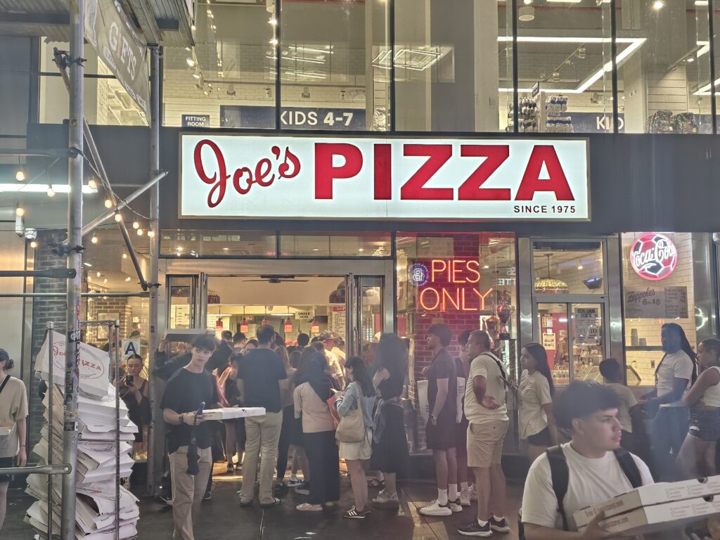 Joe's Pizza Midtown sign and customer line at night near Times Square
