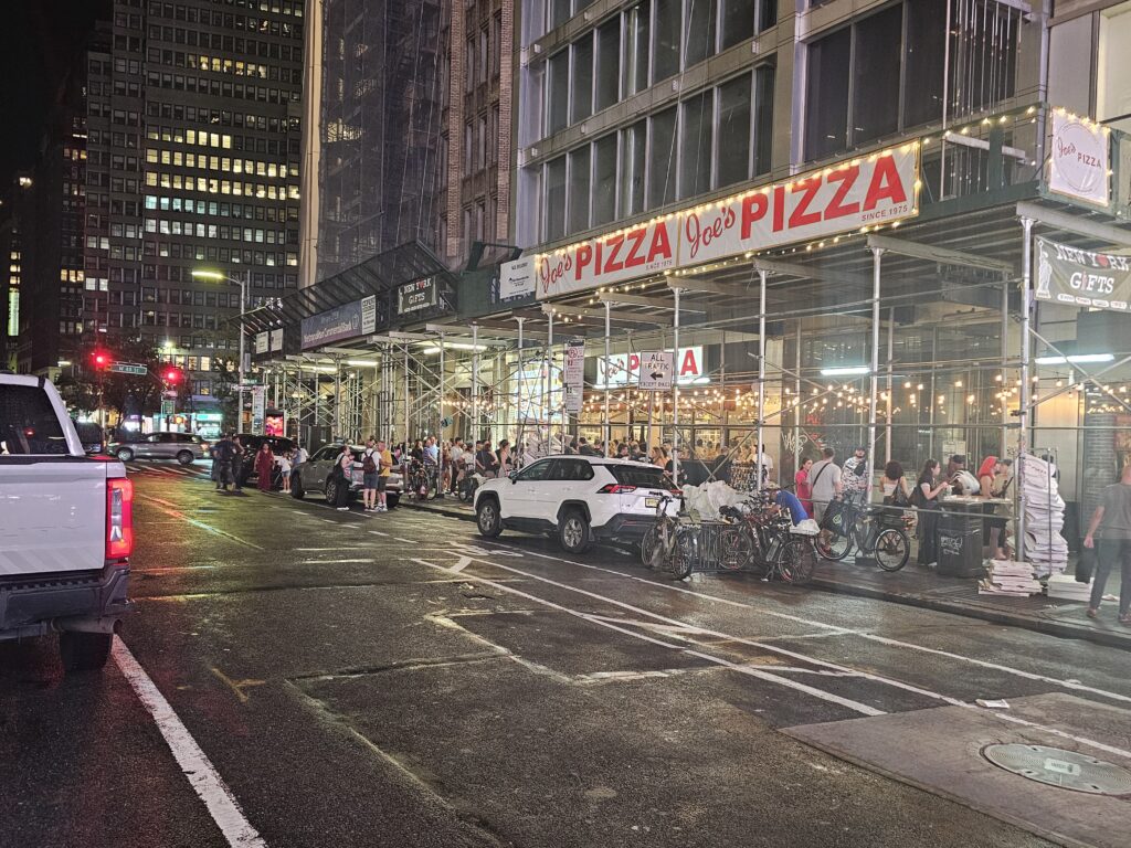 Joe's Pizza Midtown block at night with scaffolding and customers on Broadway