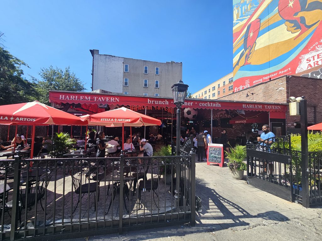 Wide street view of Harlem Tavern NYC storefront with red awnings and mural backdrop