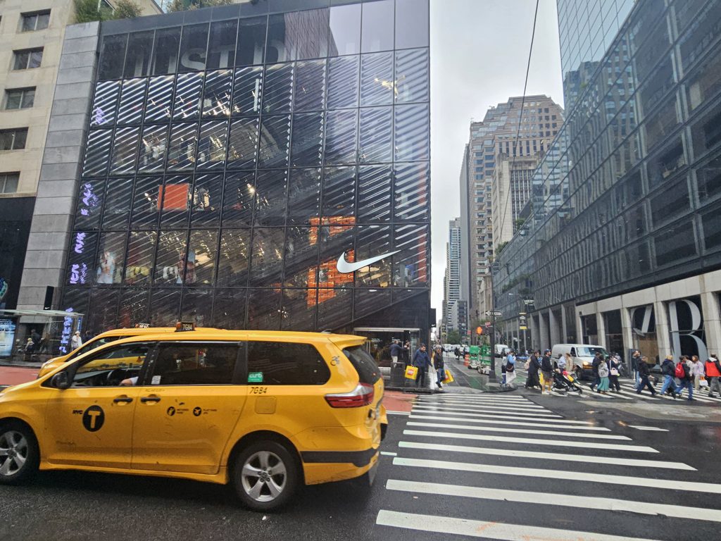 Curved slumped-glass facade of Nike House of Innovation NYC with a yellow taxi passing on 5th Avenue
